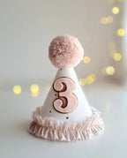White party hat with pink number 3 and pom-pom on a blurred light background.