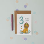 Children's birthday card with a lion illustration and 'Three Today' text, accompanied by a pen and brown envelope on a light blue background with polka dots.