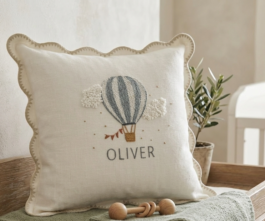Decorative pillow with embroidered hot air balloon and  text, placed on a surface with a plant in the background -  personalised baby / toddler gift.