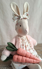 Stuffed rabbit toy with a floral bow and pink outfit holding two carrots on a wooden surface.  -   baby / toddler heirloom gift.
