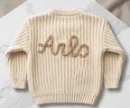 Beige knitted sweater with a name embroidered on the back, placed on a marble surface  -  personalised baby / toddler gift.