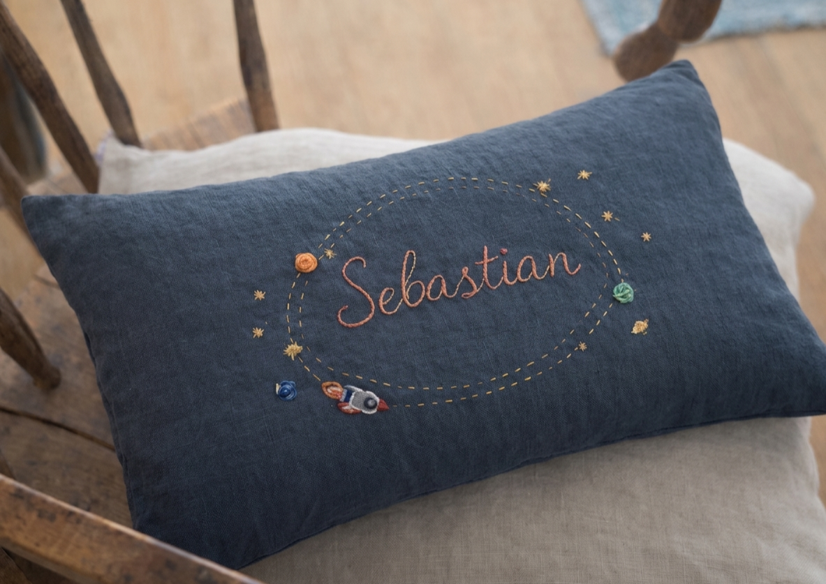 Navy blue pillow with embroidered name 'Sebastian' and space-themed decorations on a wooden chair.
